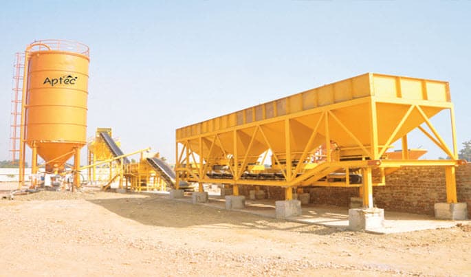 The Aptec Wet Mix Macadam plant is a stationary batching plant featuring aggregate cold feed bins, a pugmill mixing unit, and water spraying system, designed for producing well-graded, uniformly moistened granular material (WMM) used as a base/sub-base layer in road construction.